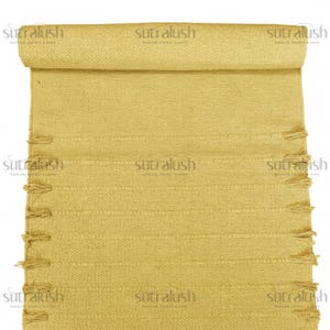 May include: A rolled-up, mustard-yellow woven rug with a textured surface and decorative fringe along the edges. The rug features horizontal lines and the text "sutralush" repeated across the top and bottom.