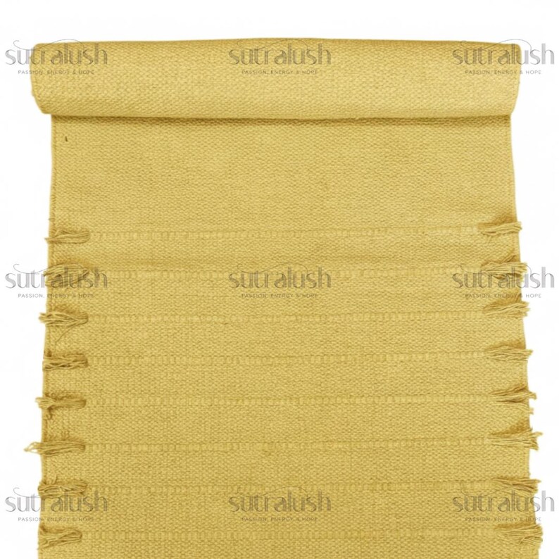 May include: A rolled-up, mustard-yellow woven rug with a textured surface and decorative fringe along the edges. The rug features horizontal lines and the text "sutralush" repeated across the top and bottom.