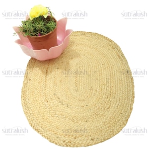 May include: A round, natural jute rug with a woven texture. A small potted plant with green foliage and a yellow flower sits in a pink, lotus-shaped planter. The rug is beige and the planter is pink.