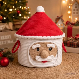 May include: A round white rope storage basket featuring a red conical lid with a white pom-pom top. The basket displays a Santa Claus face design made of tan and white rope, including a mustache, beard, and two black eyes on the front.