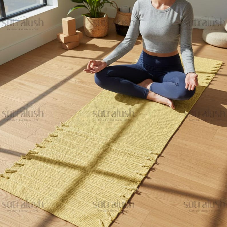 May include: A yellow yoga mat with fringe detail is laid out on a wooden floor. The mat is rectangular and has a textured surface. The image also shows a person in a gray long-sleeve shirt and blue leggings.