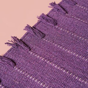 Op de afbeelding: Een dieppaars geweven textiel met een gestructureerd oppervlak met subtiele witte verticale stikdetails en een reeks kleine, bijpassende paarse franje kwastjes bevestigd langs de rechte rand van het stofmateriaal.