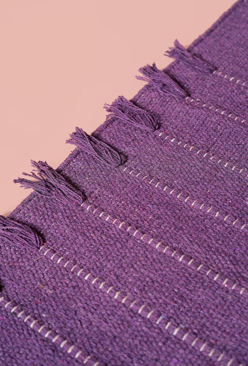 Op de afbeelding: Een dieppaars geweven textiel met een gestructureerd oppervlak met subtiele witte verticale stikdetails en een reeks kleine, bijpassende paarse franje kwastjes bevestigd langs de rechte rand van het stofmateriaal.