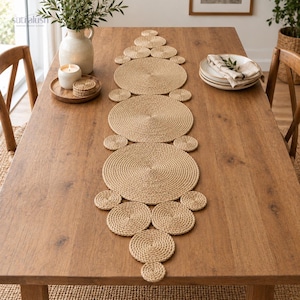 May include: A table runner composed of multiple circular woven jute discs of varying sizes arranged in a linear, symmetrical pattern. The natural tan fibre texture creates a continuous, interconnected design that spans the length of the wooden table.