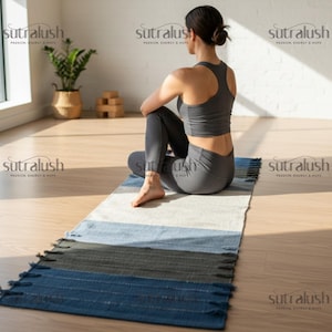 May include: A yoga mat with blue, gray, and white stripes and fringe. A person is sitting on the mat, wearing a gray sports bra and leggings. The mat is on a wooden floor, with a plant and wooden blocks in the background.