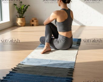 Op de afbeelding: Een gestreepte yogamat in blauwe, grijze en witte tinten, met een gefranjerde rand. Een persoon in een grijze sport-bh en legging zit op de mat. De mat ligt op een houten vloer, met een plant en blokken op de achtergrond.