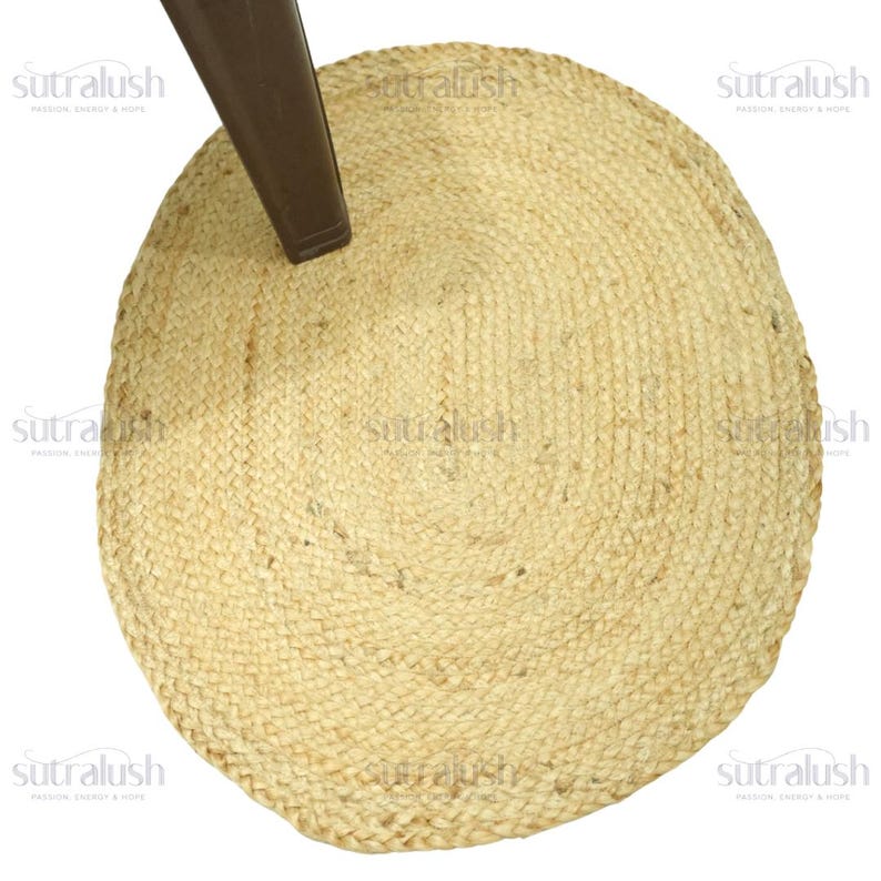 May include: A round, natural-toned jute rug with a woven texture. The rug is partially obscured by a brown chair leg. The rug's design features concentric circles, creating a textured surface. The rug is suitable for indoor use.
