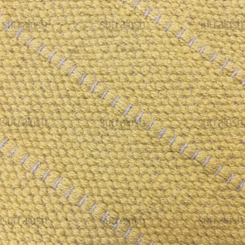 May include: Close-up of a textured yellow fabric with diagonal rows of white stitching. The fabric has a woven appearance with a slightly raised, dotted texture. The stitching is evenly spaced.