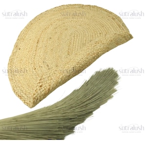 May include: A half-circle, woven jute rug in a natural beige color. The rug has a textured surface and a braided edge. A bundle of light green fibers is in the foreground, possibly a brush or decorative element.