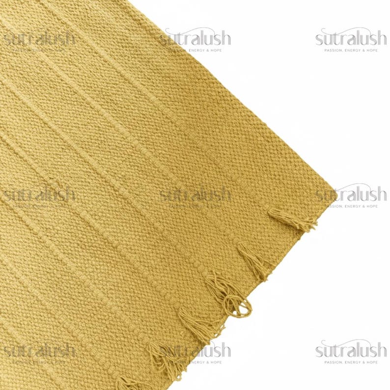 May include: Mustard yellow woven rug with vertical ribbed pattern and fringe detail. The rug's texture is visible, and the corner is shown against a white background. The rug is a home decor item.