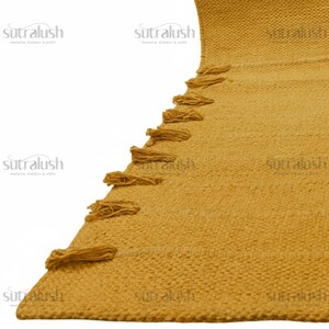 May include: Mustard yellow woven rug with a textured surface and decorative tassels along one edge. The rug has a geometric pattern and is made of a thick, durable material. The brand name "sutralush" is printed in white on the rug.