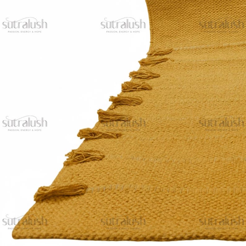 May include: Mustard yellow woven rug with a textured surface and decorative tassels along one edge. The rug has a geometric pattern and is made of a thick, durable material. The brand name "sutralush" is printed in white on the rug.