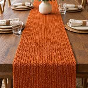 May include: A long, rectangular, burnt orange table runner featuring a thick, chunky knit texture. The runner is centered on a wooden dining table, which is set with four place settings, each including a plate, a napkin, and a glass.