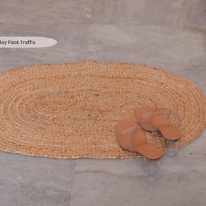 May include: Oval-shaped, woven jute rug in natural tones. The rug is paired with a pair of light brown sandals with clear straps and heels. The rug is on a gray tiled floor.