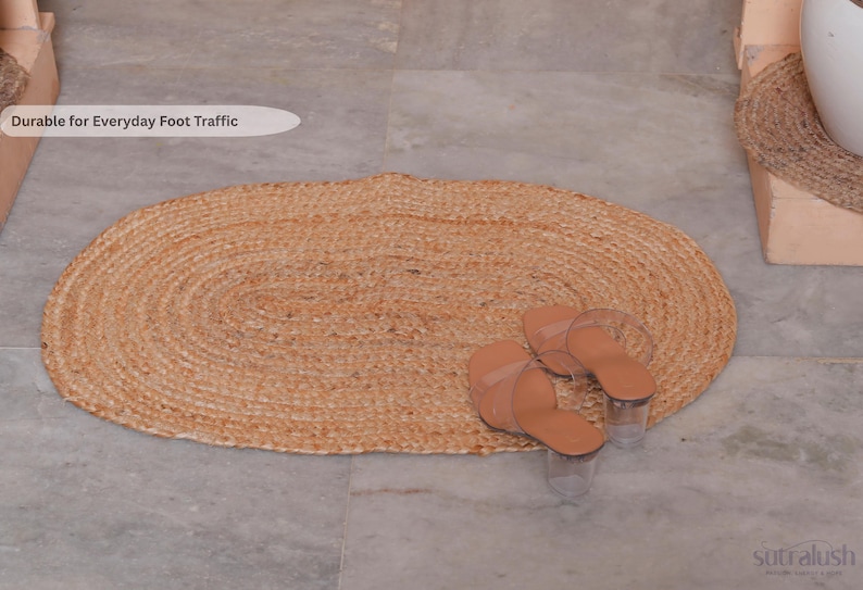May include: Oval-shaped, woven jute rug in natural tones. The rug is paired with a pair of light brown sandals with clear straps and heels. The rug is on a gray tiled floor.