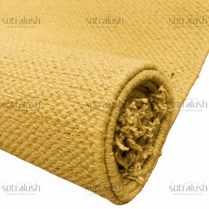 May include: A rolled-up, textured rug in a warm, golden-yellow hue. The close-up shot highlights the woven details and the fringed edge of the rug. The brand name "sutralush" is visible in the corners.