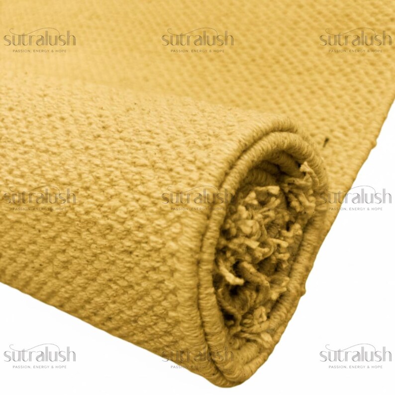 May include: A rolled-up, textured rug in a warm, golden-yellow hue. The close-up shot highlights the woven details and the fringed edge of the rug. The brand name "sutralush" is visible in the corners.
