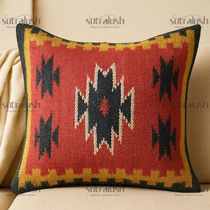 May include: A square decorative pillow with a geometric design. The pillow features a red center with black and white geometric shapes. The border is yellow and black. The pillow is on a beige sofa. The brand name "sutralush" is printed on the pillow.