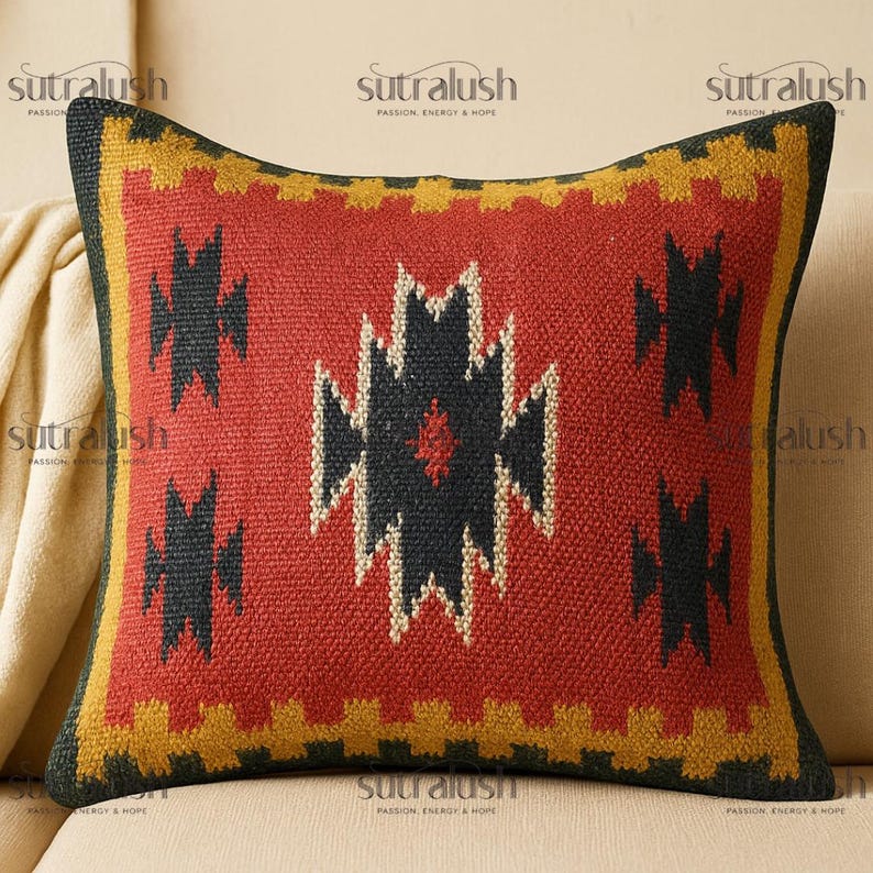May include: A square decorative pillow with a geometric design. The pillow features a red center with black and white geometric shapes. The border is yellow and black. The pillow is on a beige sofa. The brand name "sutralush" is printed on the pillow.