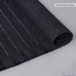 Op de afbeelding: Een zwarte geweven loper met dunne witte verticale strepen en kleine kwastjes langs de randen. De stof heeft een getextureerd uiterlijk met een label waarop 100% Natural Fiber staat. De merknaam sutralush staat in de hoek onderaan.