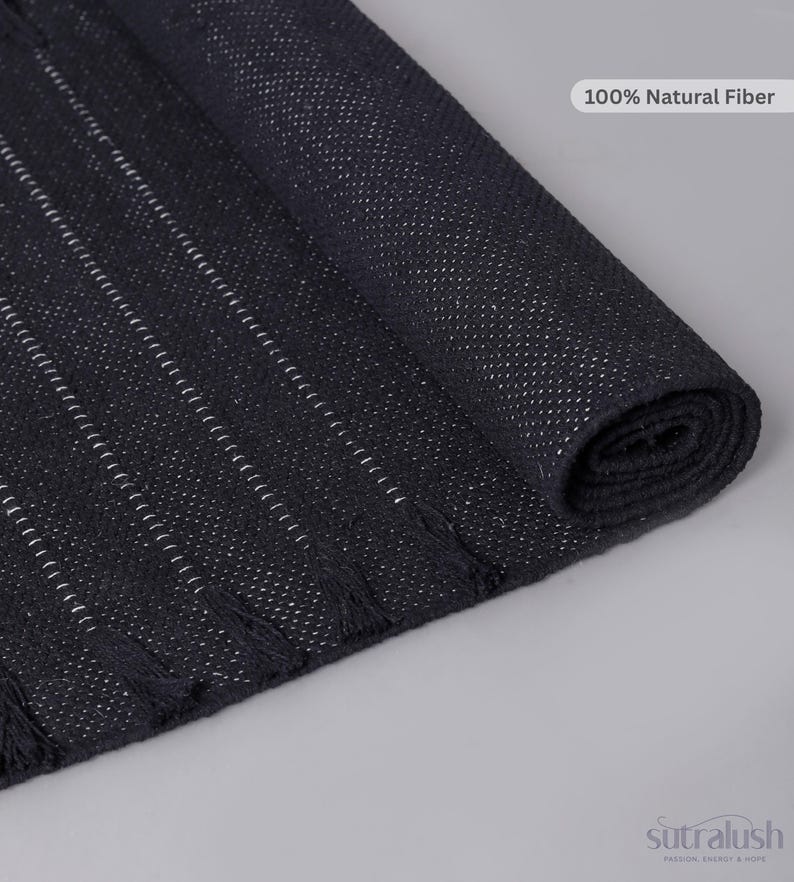 Op de afbeelding: Een zwarte geweven loper met dunne witte verticale strepen en kleine kwastjes langs de randen. De stof heeft een getextureerd uiterlijk met een label waarop 100% Natural Fiber staat. De merknaam sutralush staat in de hoek onderaan.