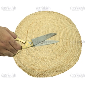 May include: A round, woven jute placemat in natural tones. A pair of gold-handled scissors is cutting the placemat. The placemat is made of tightly woven jute fibers, creating a textured surface. The scissors are open, suggesting a craft or DIY project.