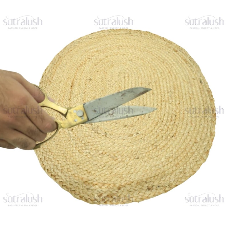 May include: A round, woven jute placemat in natural tones. A pair of gold-handled scissors is cutting the placemat. The placemat is made of tightly woven jute fibers, creating a textured surface. The scissors are open, suggesting a craft or DIY project.