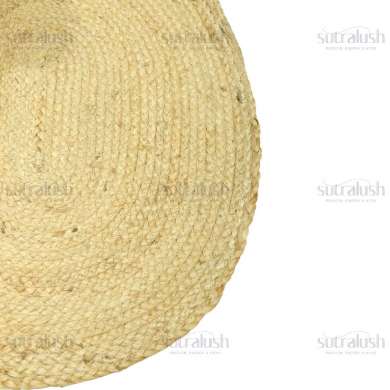 May include: A portion of a round, natural jute rug. The rug has a woven texture with a light tan color. The edge of the rug is visible, showing the circular shape. The rug is on a white background.