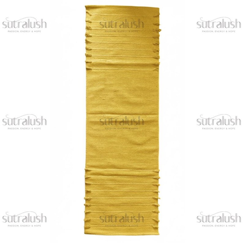 May include: A rectangular, golden-yellow runner rug with a textured surface. The rug has a fringed edge at the top and bottom. The text "sutralush" and "PASSION ENERGY & HOPE" is printed on the rug.