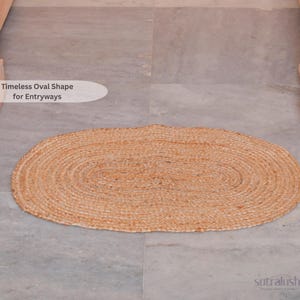 May include: An oval-shaped, woven jute rug in natural tan tones. The rug is placed on a gray tiled floor. Text on the image reads "Timeless Oval Shape for Entryways". The rug is a neutral accent piece for home decor.