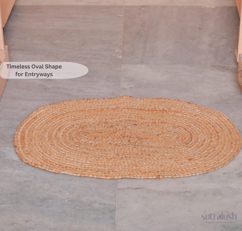 May include: An oval-shaped, woven jute rug in natural tan tones. The rug is placed on a gray tiled floor. Text on the image reads "Timeless Oval Shape for Entryways". The rug is a neutral accent piece for home decor.