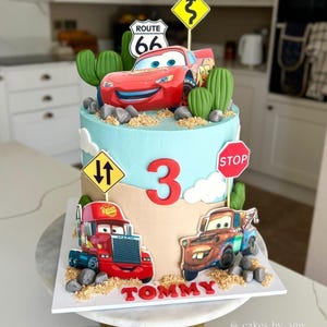 Op de afbeelding: Een cilindrische taart versierd met lichtblauw en beige glazuur, voorzien van eetbare afbeeldingen van Bliksem McQueen, Mack en Takel. De taart bevat groene cactus-toppers, verkeersborden, een rood cijfer 3 en de naam TOMMY in het rood.
