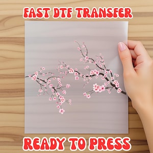 May include: A sheet featuring a floral design of pink cherry blossoms on a gray background. The sheet is held by a hand and has the text "FAST DTF TRANSFER" and "READY TO PRESS" in red.