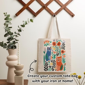 May include: A beige tote bag with a colorful floral and cat design. The illustration features a blue cat and an orange cat surrounded by flowers and foliage. The text reads: Create your custom tote bag with your iron at home!