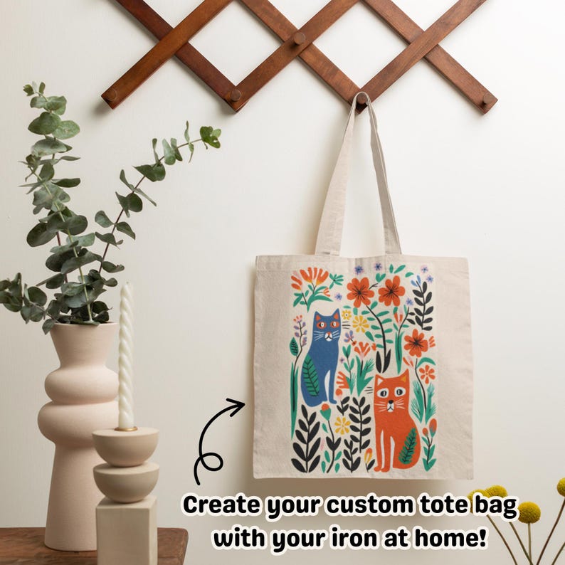 May include: A beige tote bag with a colorful floral and cat design. The illustration features a blue cat and an orange cat surrounded by flowers and foliage. The text reads: Create your custom tote bag with your iron at home!