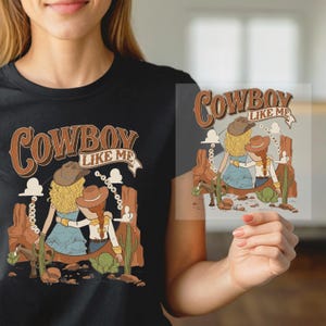 May include: A black t-shirt featuring a graphic illustration of two people wearing cowboy hats in a desert landscape with cacti and rock formations. The text "COWBOY LIKE ME" is printed in a brown, stylized font above the figures.
