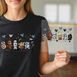 May include: Black t-shirt featuring a Star Wars-themed design. The graphic includes cartoon versions of characters like R2-D2, Darth Vader, and Yoda, along with decorative elements. The design is repeated on a transparent sheet.