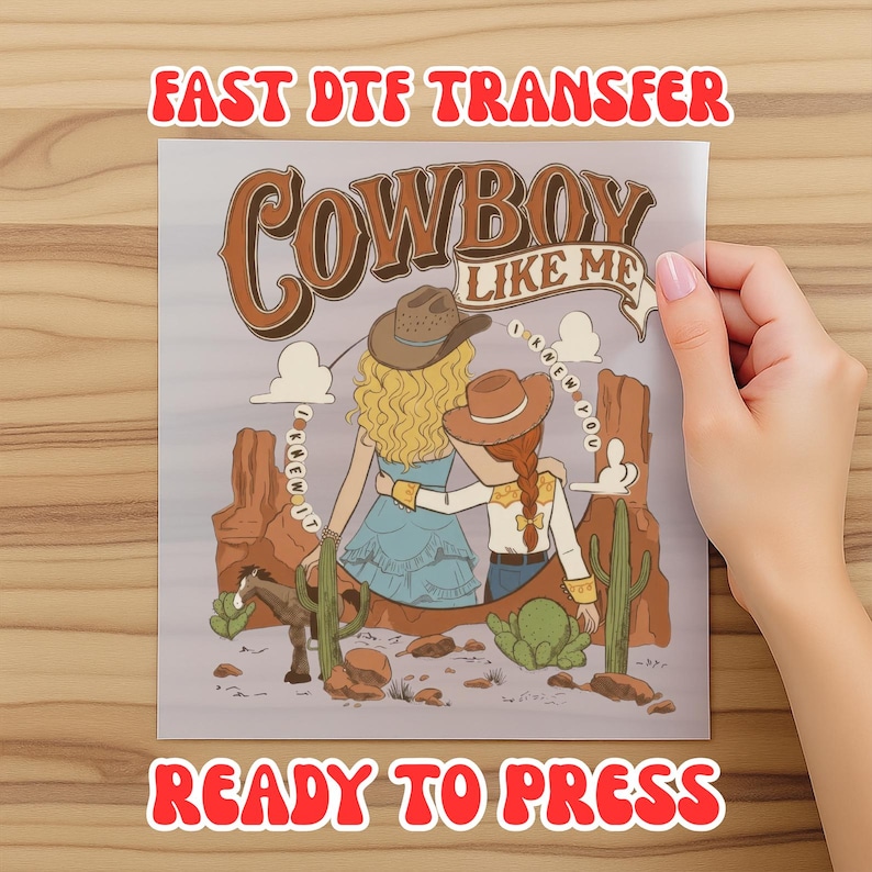 May include: A DTF transfer sheet featuring a desert scene with two figures wearing cowboy hats, one with blonde hair and one with red hair, standing before rock formations and cacti. The text reads COWBOY LIKE ME and I KNEW IT.