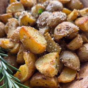 Könnte beinhalten: Eine Schüssel mit gerösteten Kartoffelspalten, gewürzt mit Kräutern und bestreut mit geriebenem Käse. Die goldbraunen Kartoffeln sind in kleine Stücke geschnitten und mit frischen grünen Rosmarinzweigen garniert, serviert in einer rustikalen Holzschale.