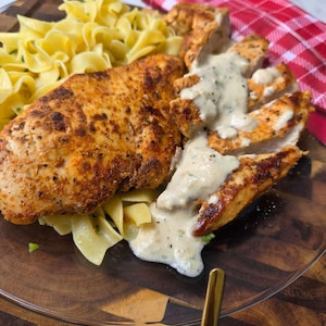 May include: A plate of seasoned, pan-seared chicken breast, with one portion sliced and topped with a creamy white herb sauce, served alongside a generous portion of yellow egg noodles. The meal is presented on a clear glass plate.