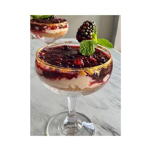 May include: A dessert in a clear glass with a gold rim, layered with a crumbly base, a creamy white layer, and a dark berry topping. Garnished with a blackberry and mint leaves. Another glass is visible in the background.