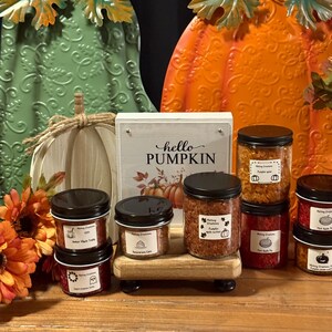 May include: A fall-themed display featuring decorative pumpkins and jars of scented wax melts. The sign reads "hello PUMPKIN". The jars are filled with colorful wax melts and have black lids. The pumpkins are green, white, and orange.