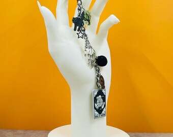 May include: A silver keychain with various charms, including a black cat, a star, a skeleton hand, a black sphere, and a tarot card with the words "WHEEL OF FORTUNE". The keychain is displayed on a white hand-shaped stand against an orange background.