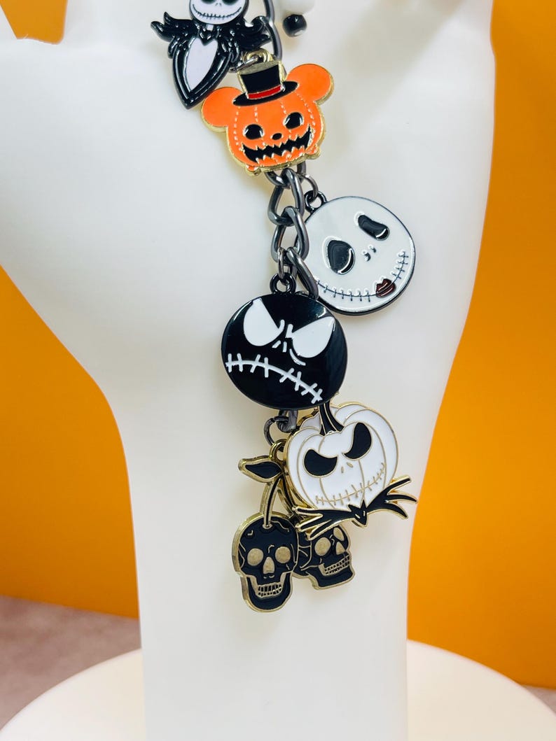 Puede incluir: Una pulsera de dijes con colgantes tem&aacute;ticos de Halloween. Los dijes incluyen a Jack Skellington, una calabaza con sombrero de copa y dise&ntilde;os de calaveras. Los dijes son negros, blancos y naranjas con detalles dorados, unidos por una cadena de metal oscuro.