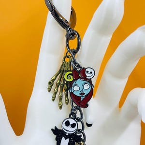 Puede incluir: Un llavero tem&aacute;tico de Pesadilla antes de Navidad con m&uacute;ltiples dijes. Los dijes incluyen a Sally, Jack Skellington, una mano de esqueleto y una calabaza. El llavero est&aacute; sujeto a un cierre de metal negro.