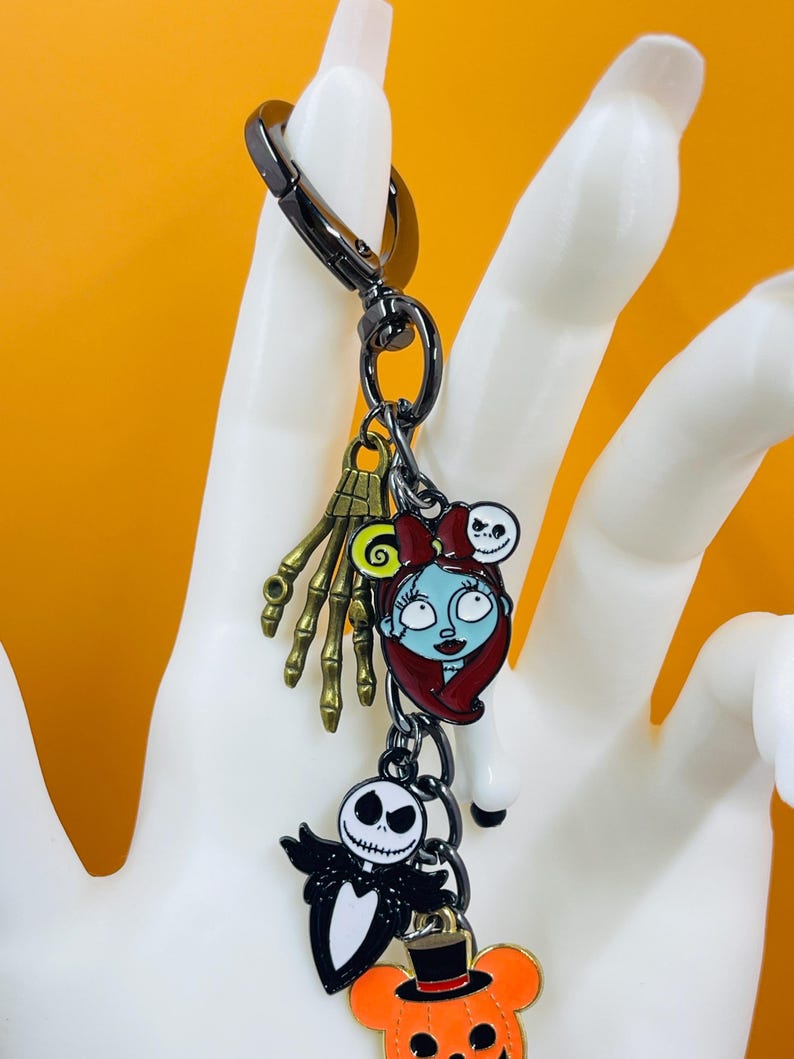 Puede incluir: Un llavero tem&aacute;tico de Pesadilla antes de Navidad con m&uacute;ltiples dijes. Los dijes incluyen a Sally, Jack Skellington, una mano de esqueleto y una calabaza. El llavero est&aacute; sujeto a un cierre de metal negro.
