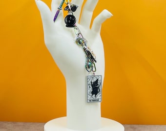 May include: A keychain with a rainbow-colored crescent moon, sword, and various charms. A black crystal ball and a tarot card with a cat illustration are also attached. The keychain is displayed on a white hand-shaped stand against an orange background.