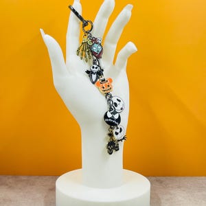 Puede incluir: Un llavero tem&aacute;tico de Halloween con varios dijes de personajes. Los dijes incluyen una mano de esqueleto, un personaje con la boca cosida, un murci&eacute;lago, una calabaza y una calavera. El llavero se exhibe sobre un soporte blanco en forma de mano.