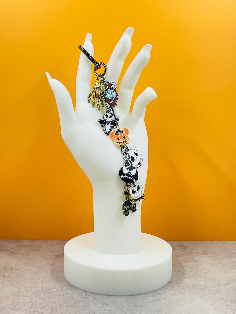 Puede incluir: Un llavero tem&aacute;tico de Halloween con varios dijes de personajes. Los dijes incluyen una mano de esqueleto, un personaje con la boca cosida, un murci&eacute;lago, una calabaza y una calavera. El llavero se exhibe sobre un soporte blanco en forma de mano.