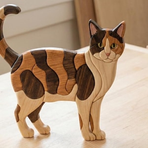 May include: A wooden sculpture of a calico cat. The cat is crafted from various shades of wood, including light, medium, and dark browns. The cat stands with its tail curved upwards. The cat's face features a light brown nose and green eyes. The sculpture rests on a light brown wooden surface.